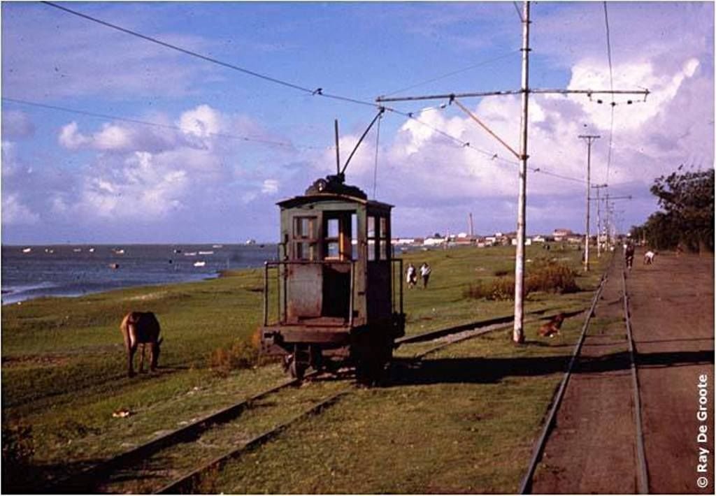 Foto de um bondinho sobre trilhos. À direita dele, grama verde, um cavalo pastando e após a Lagoa dos Patos, à esquerda uma estrada de areia. O céu está azul, com poucas núvens.