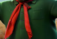 Tu sabes o que significa as cores dos lenços gaúchos? Desenho de um close-up do torso de uma figura humana. O foco principal é a roupa da figura, que consiste em uma camisa verde escura com mangas compridas. A camisa é abotoada com três botões pretos no centro, e há um bolso quadrado simples no lado direito do peito. Um lenço vermelho vibrante está amarrado ao redor do pescoço da figura.