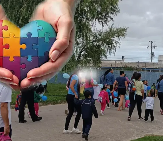 Pais, professores e alunos promovem manifestação em defesa da inclusão no bairro Nossa Senhora de Fátima A imagem mostra um grupo de adultos e crianças caminhando juntos por uma calçada, em um evento ao ar livre. Muitas crianças carregam bexigas azuis e usam mochilas escolares. Algumas pessoas vestem camisetas azuis, sugerindo uma possível campanha de conscientização. Ao fundo, há um muro azul, árvores e postes elétricos sob um céu nublado. No canto superior esquerdo da imagem, há uma montagem gráfica de duas mãos segurando um coração colorido formado por peças de quebra-cabeça — símbolo amplamente associado ao autismo.