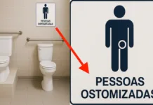 Depois de Anos de Espera, Rio Grande Inaugura Banheiro para Pessoas Ostomizadas A imagem mostra um banheiro acessível com dois vasos sanitários lado a lado, barras de apoio nas paredes e um dos vasos instalado sobre uma pequena plataforma, com apoio lateral acolchoado. Acima dos vasos, há uma placa fixada na parede com o símbolo de uma pessoa estilizada. Esse símbolo tem um círculo na altura do abdômen, representando uma bolsa de ostomia. Abaixo do desenho está escrito: “PESSOAS OSTOMIZADAS”. À direita da imagem, há um destaque ampliado da placa, com uma seta vermelha apontando para o símbolo e o texto.