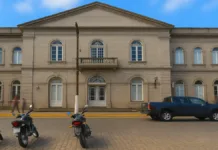 A História da Santa Casa de Rio Grande-RS: Tradição, Curiosidades e Desafios Fachada da Santa Casa de Misericórdia de Rio Grande, um prédio histórico de estilo neoclássico, construído em alvenaria de pedra com acabamento em tom cinza. A edificação tem dois andares e janelas em arco com molduras detalhadas. No centro, há uma sacada com grade de pedra acima da entrada principal, que possui uma porta com arco e vitrais. À frente do prédio, na calçada de paralelepípedos, estão estacionadas três motocicletas e uma caminhonete azul escura. Algumas pessoas transitam pela calçada, e há palmeiras visíveis ao lado esquerdo da imagem, sob um céu parcialmente nublado.