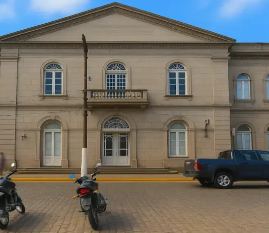 A História da Santa Casa de Rio Grande-RS: Tradição, Curiosidades e Desafios Fachada da Santa Casa de Misericórdia de Rio Grande, um prédio histórico de estilo neoclássico, construído em alvenaria de pedra com acabamento em tom cinza. A edificação tem dois andares e janelas em arco com molduras detalhadas. No centro, há uma sacada com grade de pedra acima da entrada principal, que possui uma porta com arco e vitrais. À frente do prédio, na calçada de paralelepípedos, estão estacionadas três motocicletas e uma caminhonete azul escura. Algumas pessoas transitam pela calçada, e há palmeiras visíveis ao lado esquerdo da imagem, sob um céu parcialmente nublado.