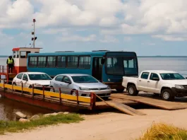 Uma balsa, um tipo de embarcação usada para transportar veículos e passageiros sobre um rio. A balsa está ancorada perto de uma margem, e três veículos estão estacionados em sua plataforma: um carro de cor branca, um carro prata e uma caminhonete também branca. Na parte superior da balsa, um homem está em uma cabine de comando, usando um colete de segurança amarelo e observando ao longe. Ao fundo, é possível ver uma vista tranquila do rio e um céu azul com algumas nuvens. A margem da balsa é composta por uma pequena área de terra com gramado e pedras visíveis.