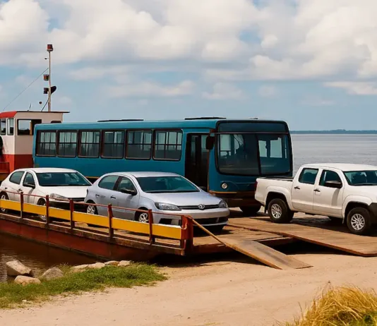 Serviço de Balsa na Ilha dos Marinheiros é Retomado Após Manutenção Técnica Uma balsa, um tipo de embarcação usada para transportar veículos e passageiros sobre um rio. A balsa está ancorada perto de uma margem, e três veículos estão estacionados em sua plataforma: um carro de cor branca, um carro prata e uma caminhonete também branca. Na parte superior da balsa, um homem está em uma cabine de comando, usando um colete de segurança amarelo e observando ao longe. Ao fundo, é possível ver uma vista tranquila do rio e um céu azul com algumas nuvens. A margem da balsa é composta por uma pequena área de terra com gramado e pedras visíveis.
